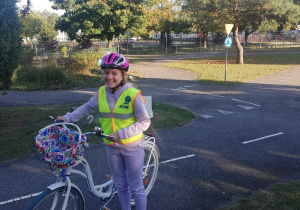 Uczennica w kamizelce odblaskowej i różowym kasku ze swoim rowerem. W tle zielony teren miasteczka ruchu drogowego.